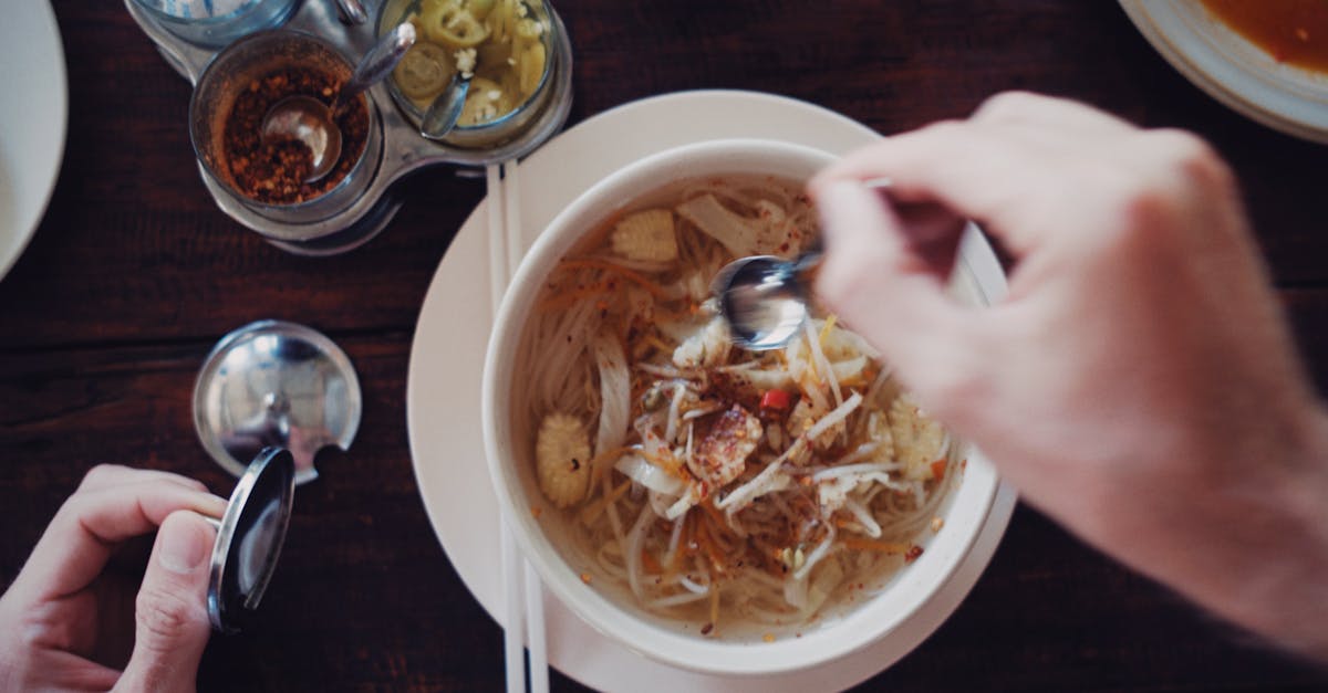 Top-view shot of hands preparing to eat a flavorful Asian noodle soup with various spices and chopsticks.