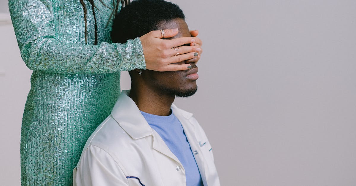 A woman in a sparkling green dress surprises a man by covering his eyes indoors.