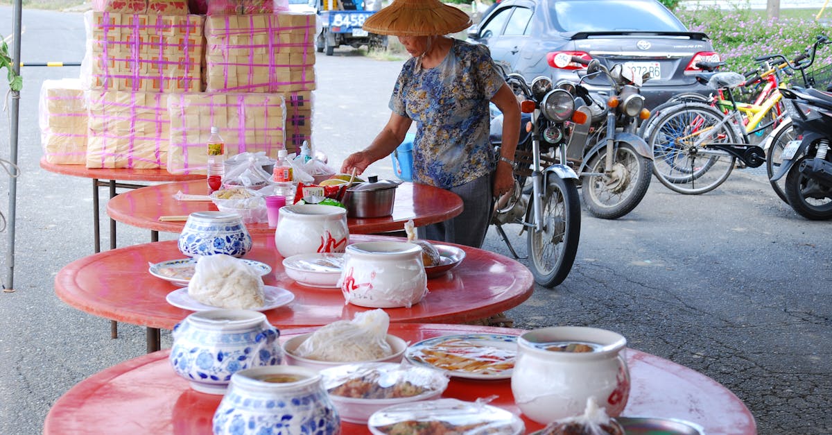 Street vendor in straw hat arranges local Asian food on red tables in outdoor market.