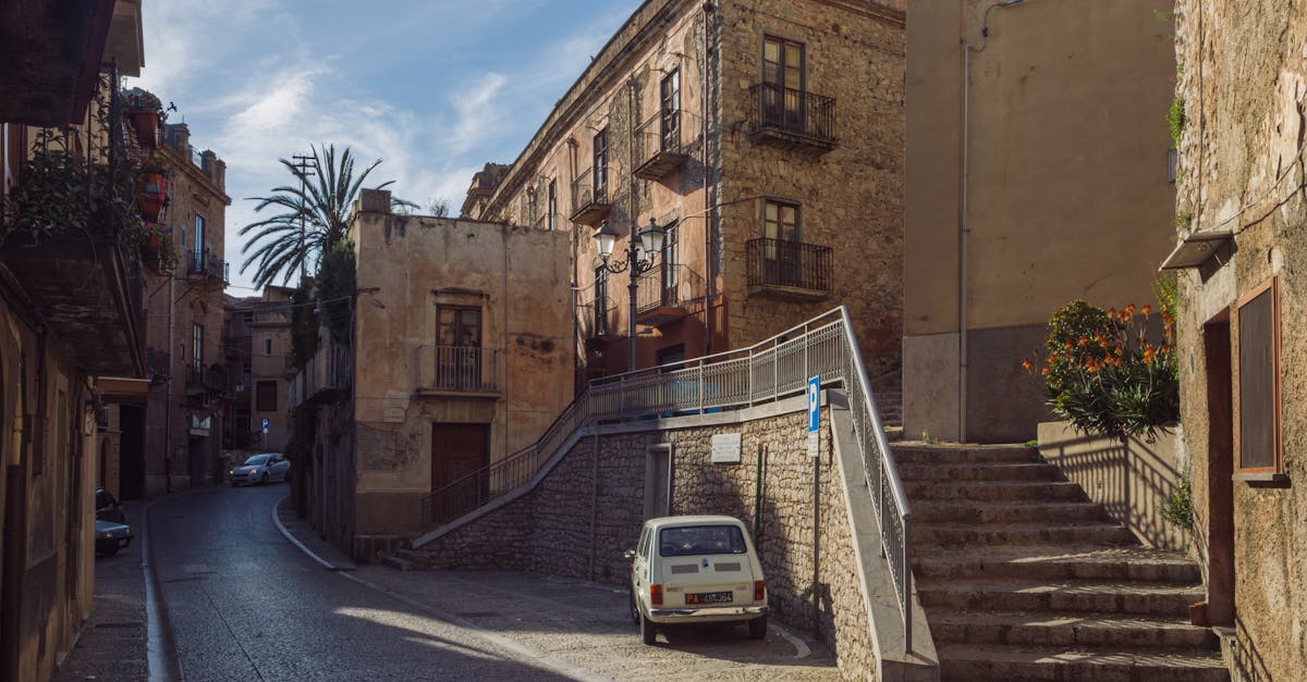 Sunlit street view in Caccamo, Sicily with classic architecture and cars.