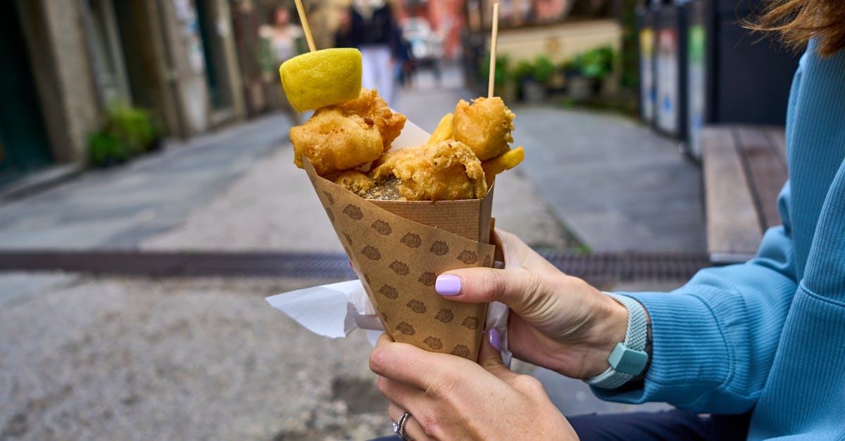 Person holding fried snacks in a paper cone outdoors on a street.