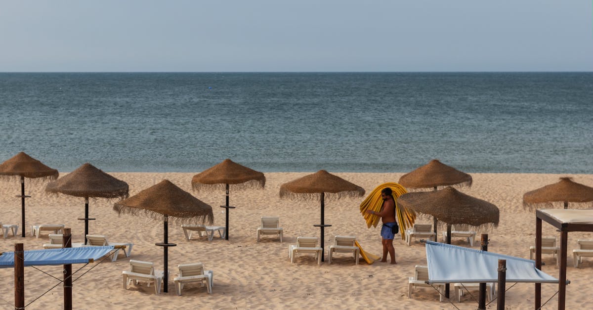 A tranquil beach scene with straw umbrellas and a person preparing sun loungers by the sea.