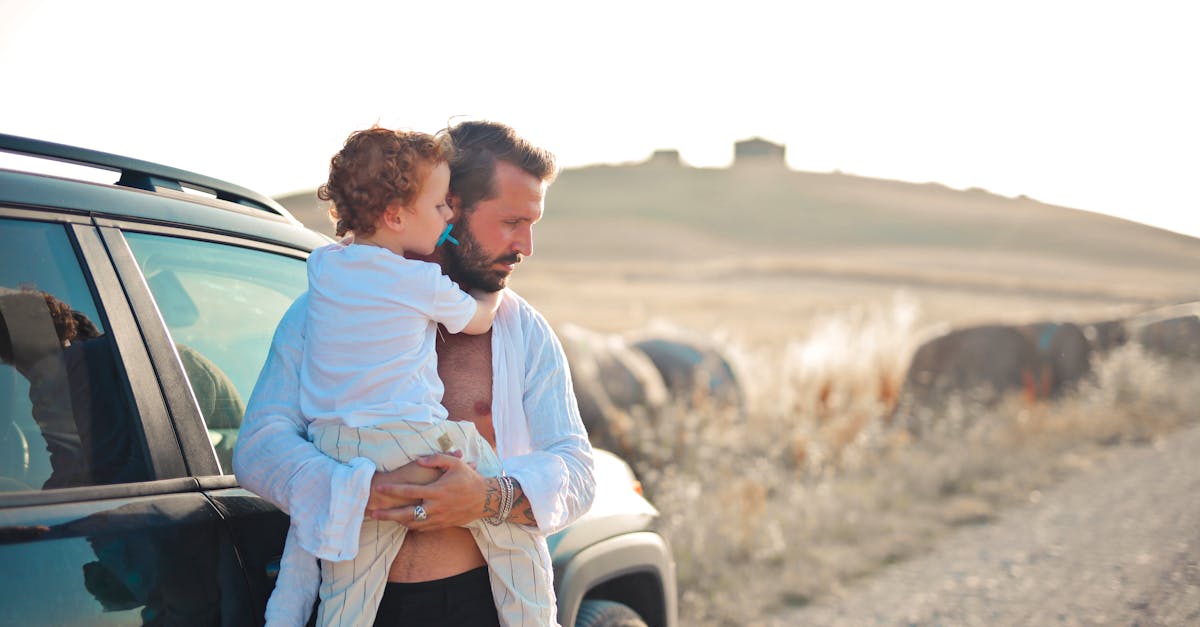 A father lovingly holds his son by a car in a peaceful countryside setting.