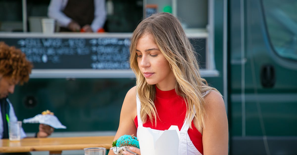 Tranquil woman with blond hair in casual clothes sitting at table with drink and eating blue burger in street cafe against food truck in daytime
