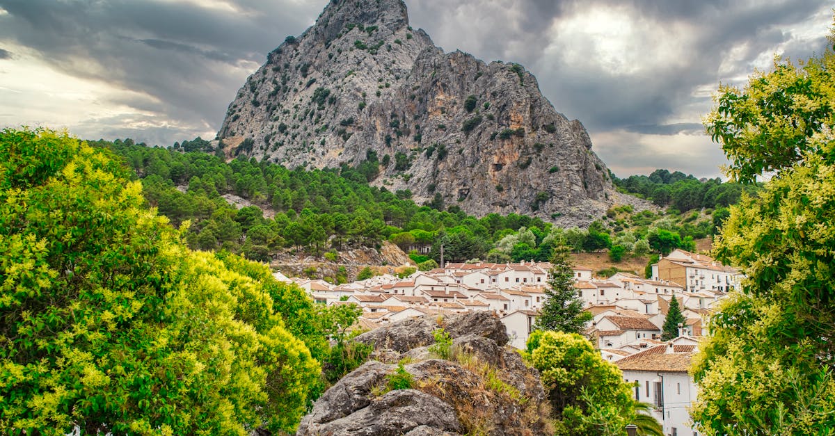 A scenic view of a small village nestled beneath a towering mountain with a dramatic cloudy sky.