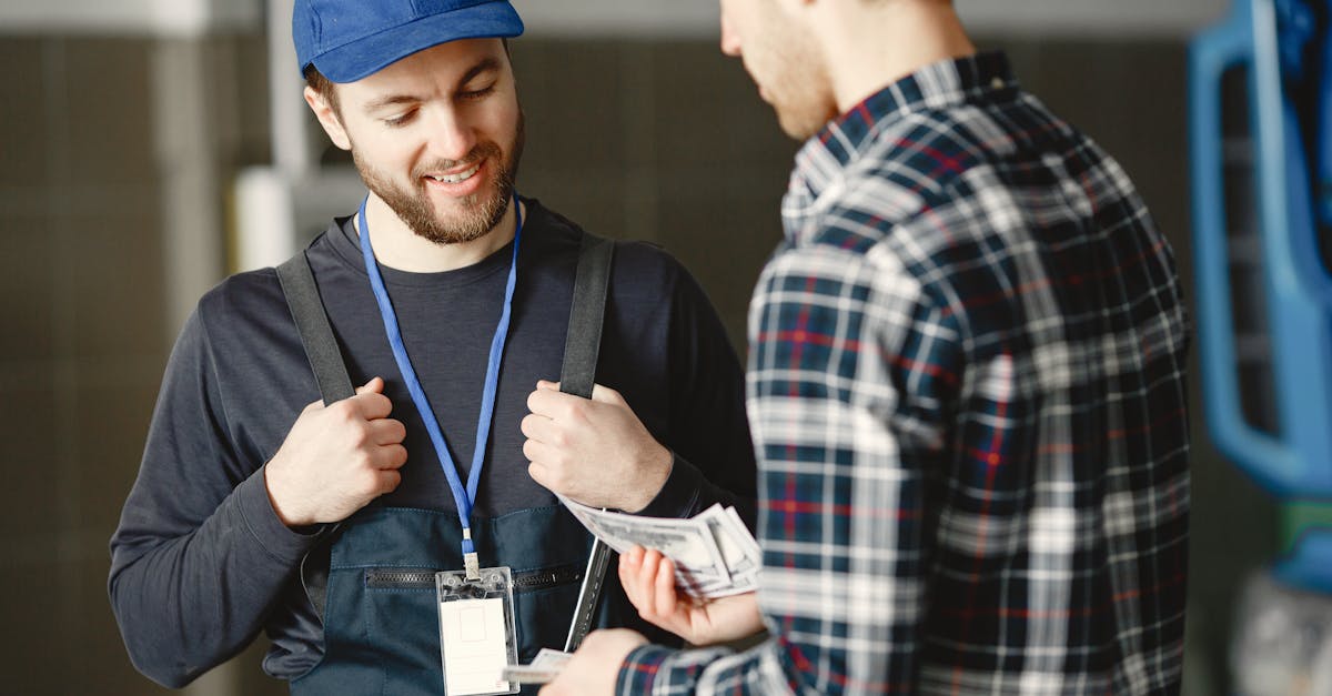 A mechanic in a blue cap receives cash payment from a customer in a plaid shirt, smiling.
