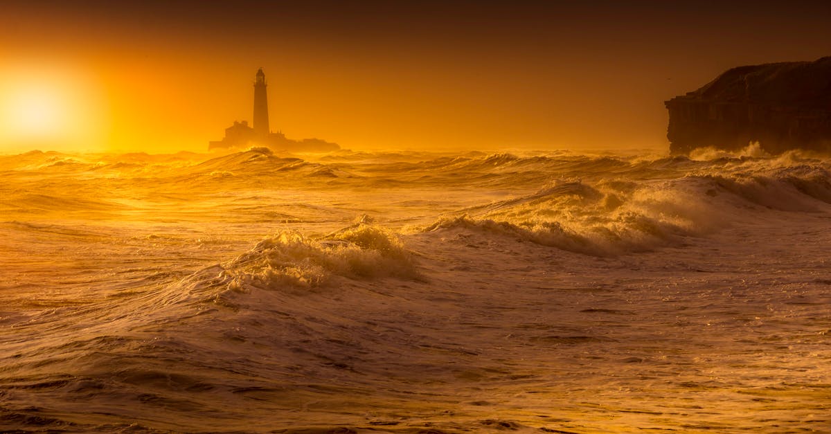 Dramatic ocean waves at sunset with a silhouetted lighthouse in view.